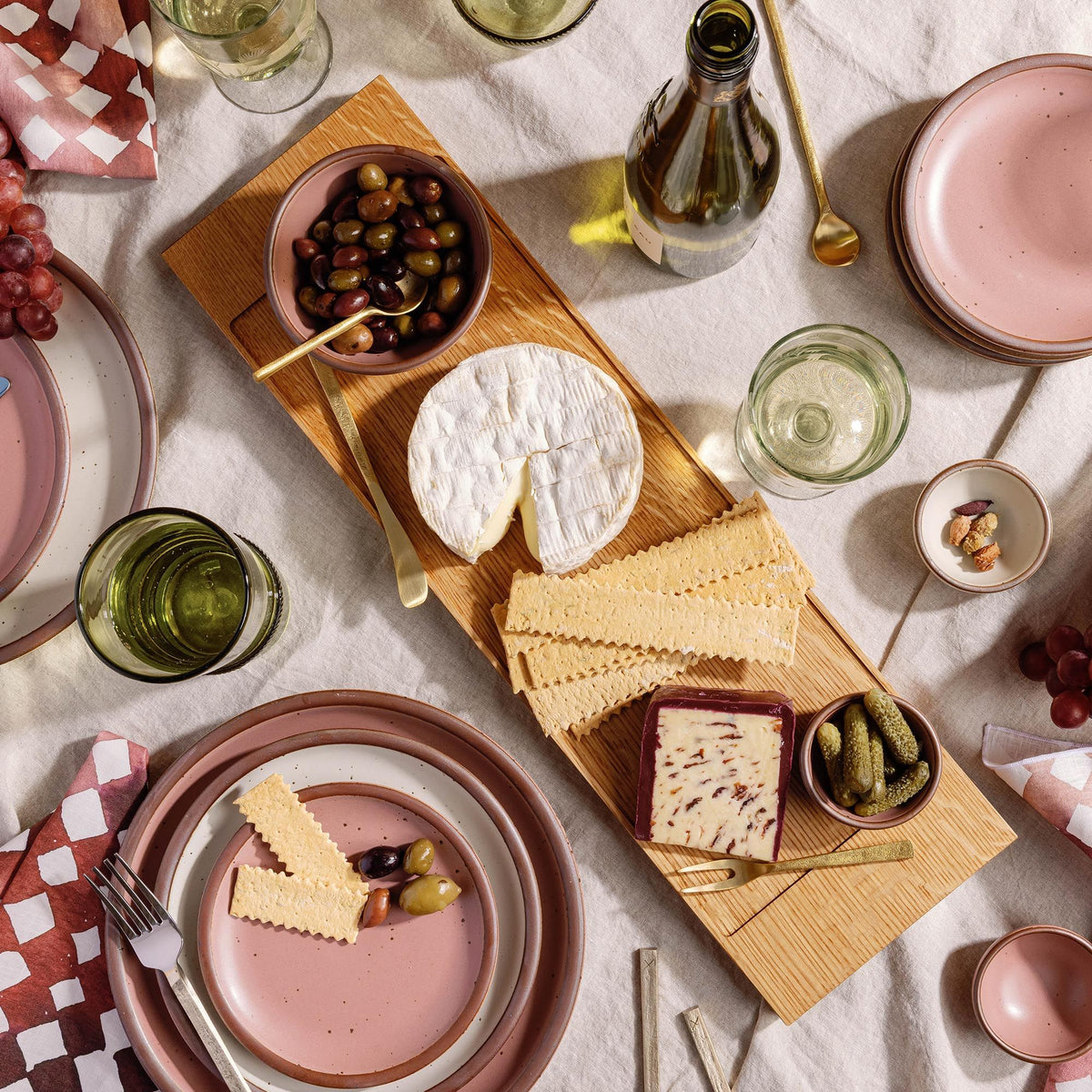 Light wood cheese board with olives, crackers, brie, pickles, and wine glasses on a linen table, surrounded by pink plates and utensils.