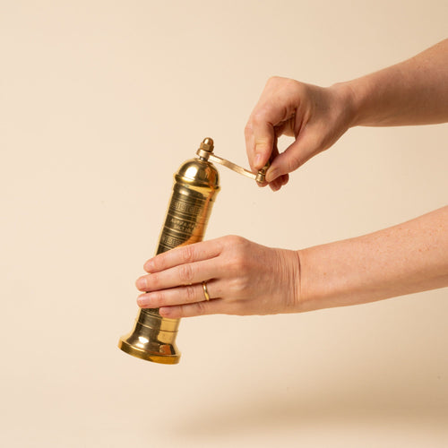A hand holds a small brass salt mill with engraved details and a side handle on a neutral background.