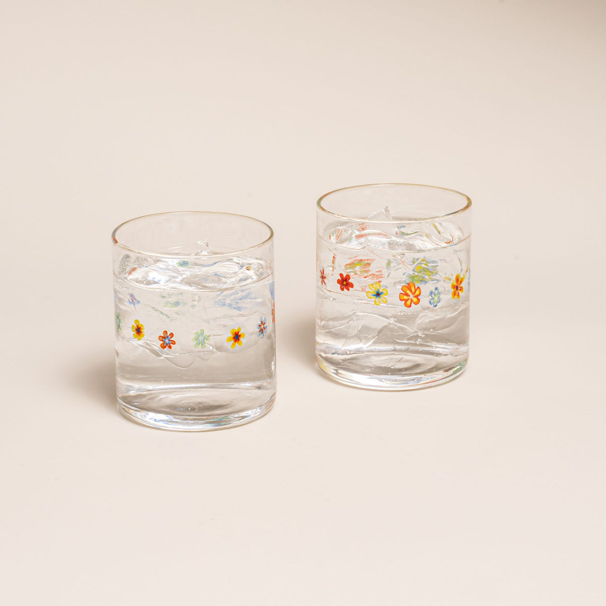 Two clear glasses with a ring of multicolored little flowers, filled with water, on a cream background.
