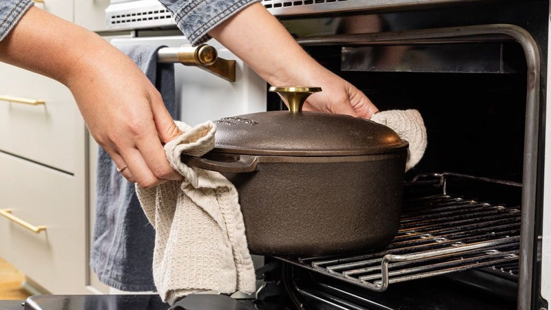 Hands hold a kitchen towel to carry a cast iron dutch oven with a handle on each side and cast iron lid out of an oven.