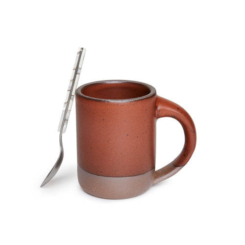 A spoon leaning on a medium sized ceramic mug with handle in a cool burnt terracotta color featuring iron speckles and unglazed rim and bottom base.