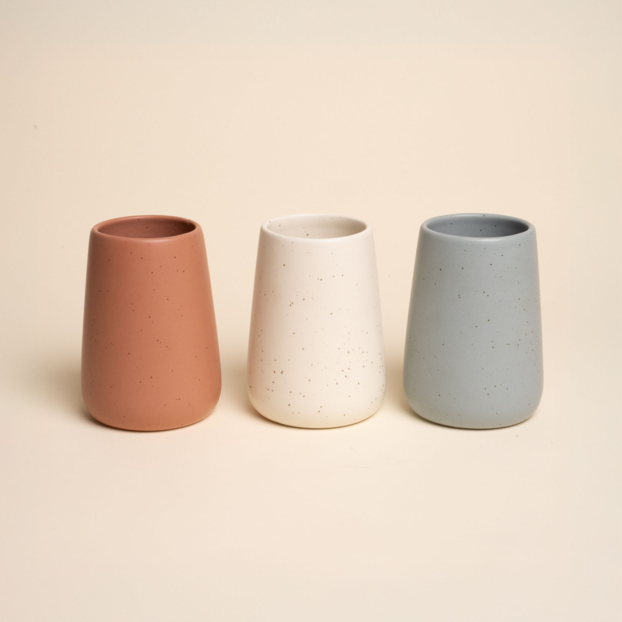 Three minimal vases in light terracotta, cream, and blue-grey tones, arranged side by side on a neutral background.