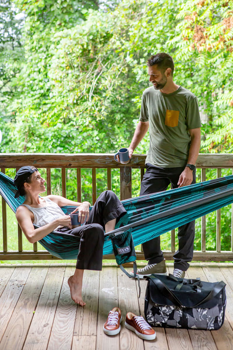 A person relaxes in a blue-green hammock on a wooden deck, holding a mug, while chatting with another person standing beside them with a mug as well. Nearby, a pair of sneakers and a black duffel bag rest on the deck, with lush greenery in the background.