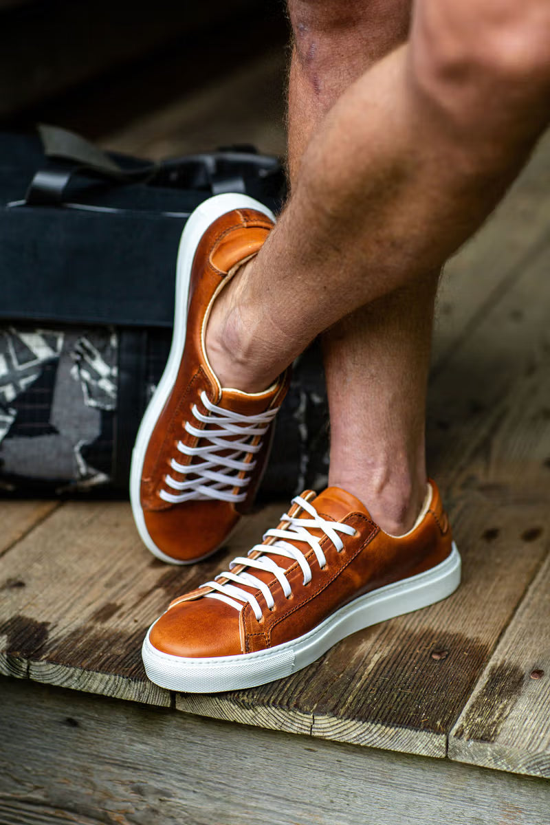 A person wearing brown leather sneakers with white laces, standing on a wooden surface next to a black bag.