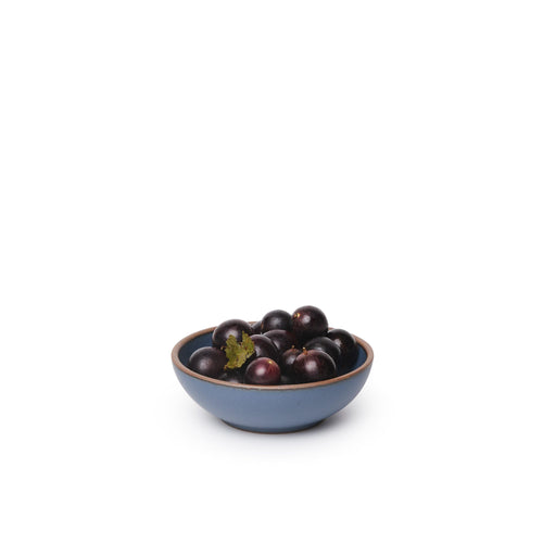 A small shallow ceramic bowl in a cool, medium blue color featuring iron speckles and an unglazed rim, filled with cherries