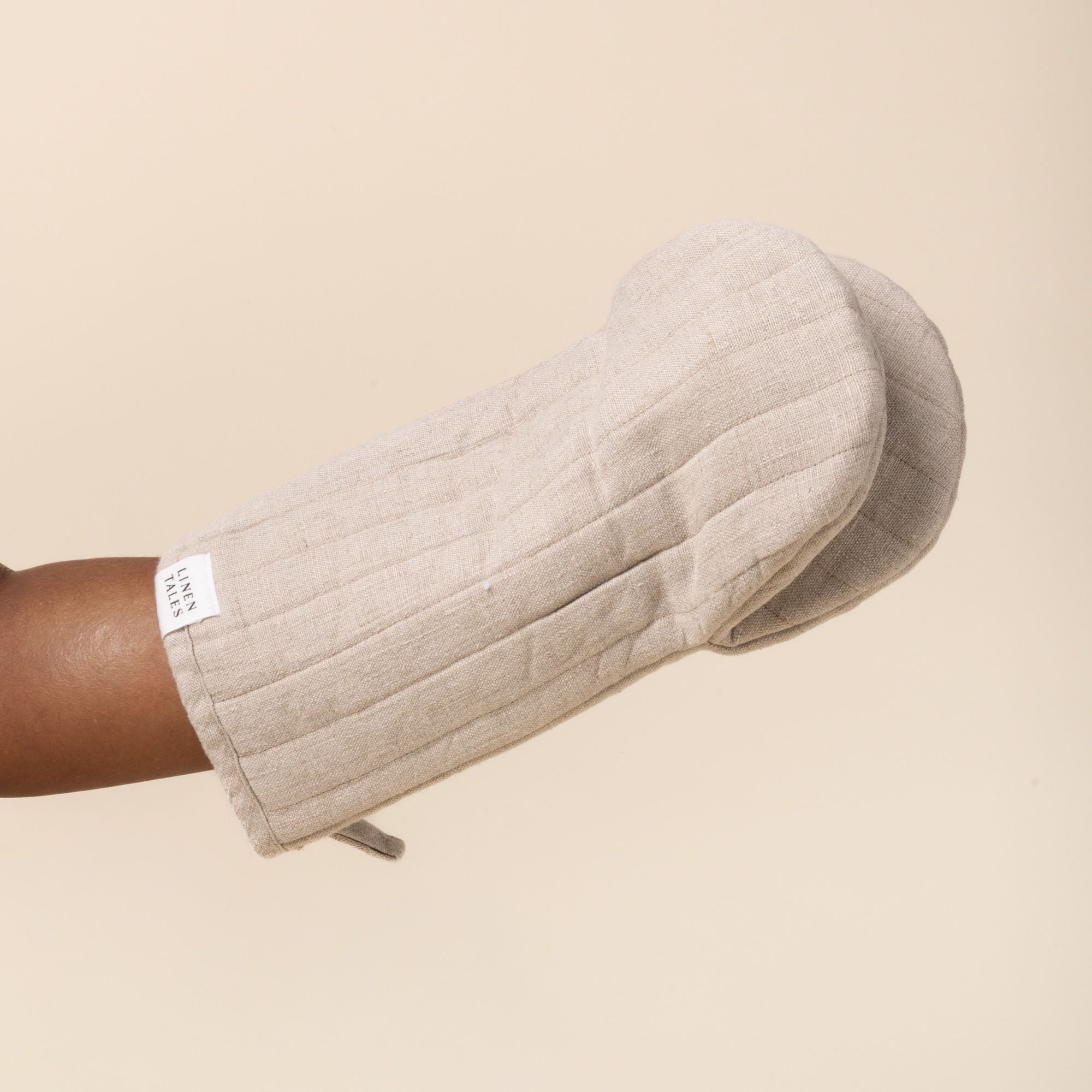 Hand inside a natural color linen oven mitt with stitched channels and a small ‘Linen Tales’ label, against a light background.