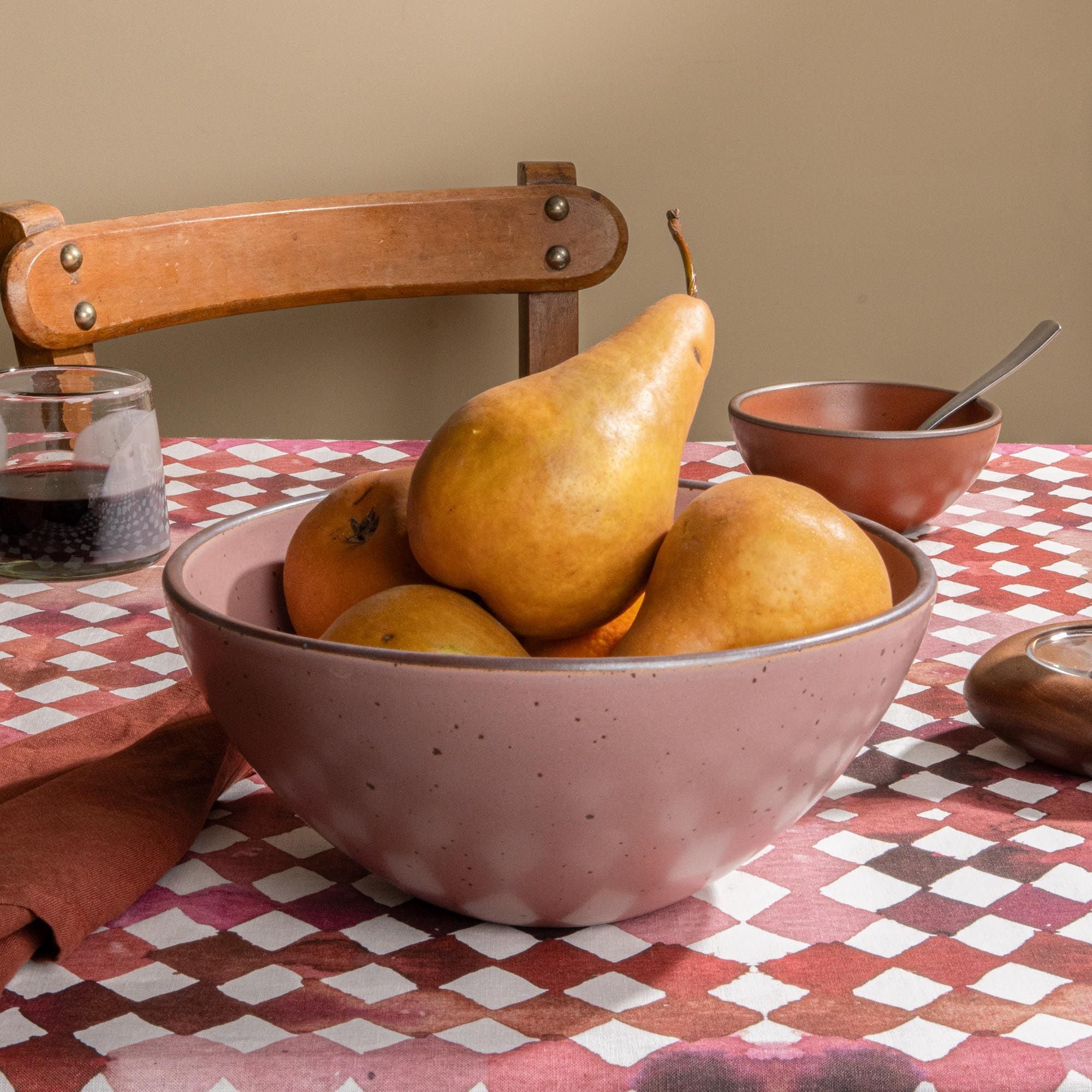 Dusty mauve pink ceramic fruit bowl filled with pears and citrus on a patterned tablecloth with a wooden chair in the background.