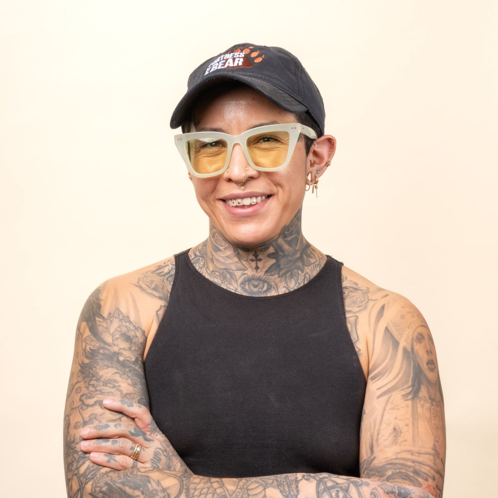 A person with tattoos wearing a black sleeveless shirt, light-colored eyeglasses, and a black cap stands against a light background, smiling with arms crossed.