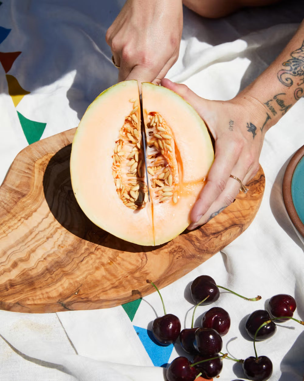 A person slices a cantaloupe in half on a wooden board, surrounded by fresh cherries on a colorful, patterned cloth.