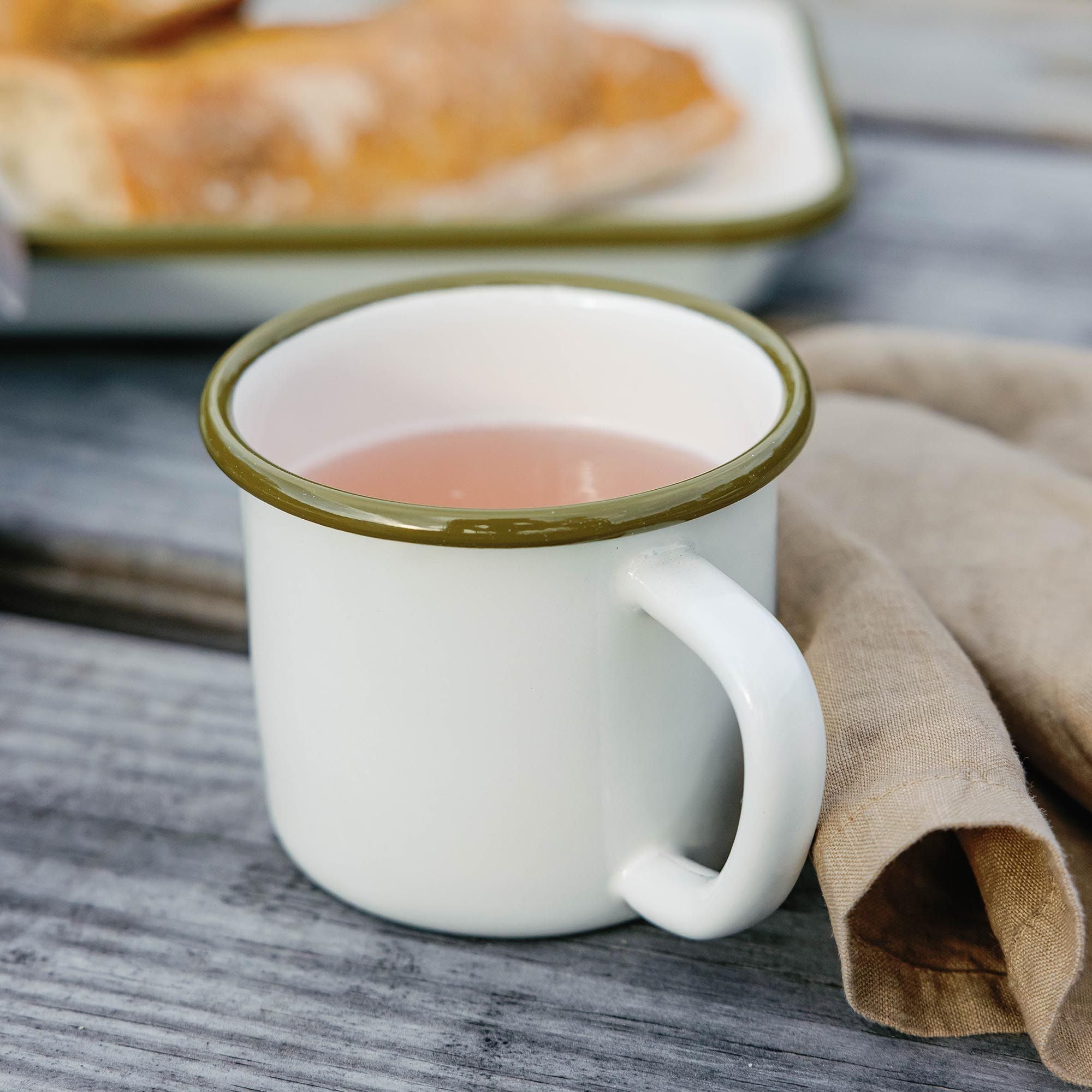 White enamel mug with a green rim filled with tea on a wooden deck, napkin and baking dish in background.