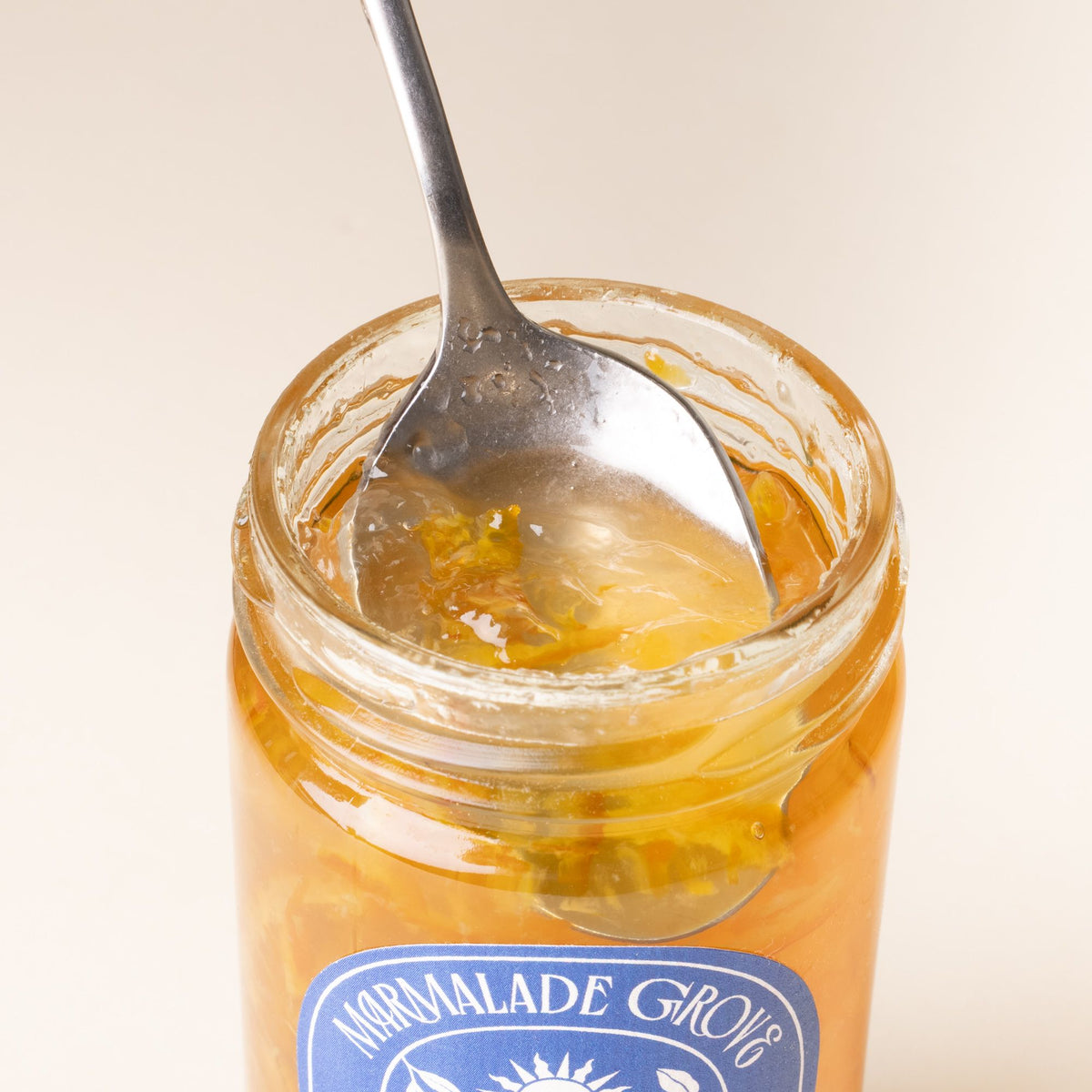 A spoon scoops marmalade out of an open jar of Marmalade Grove Pixie Tangerine, featuring a blue and white label with fruit and floral illustrations.