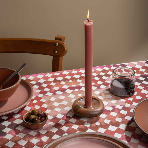 Lit pink taper candle in a wooden holder on a red and pink watercolor diamond tablecloth with tableware and olives.