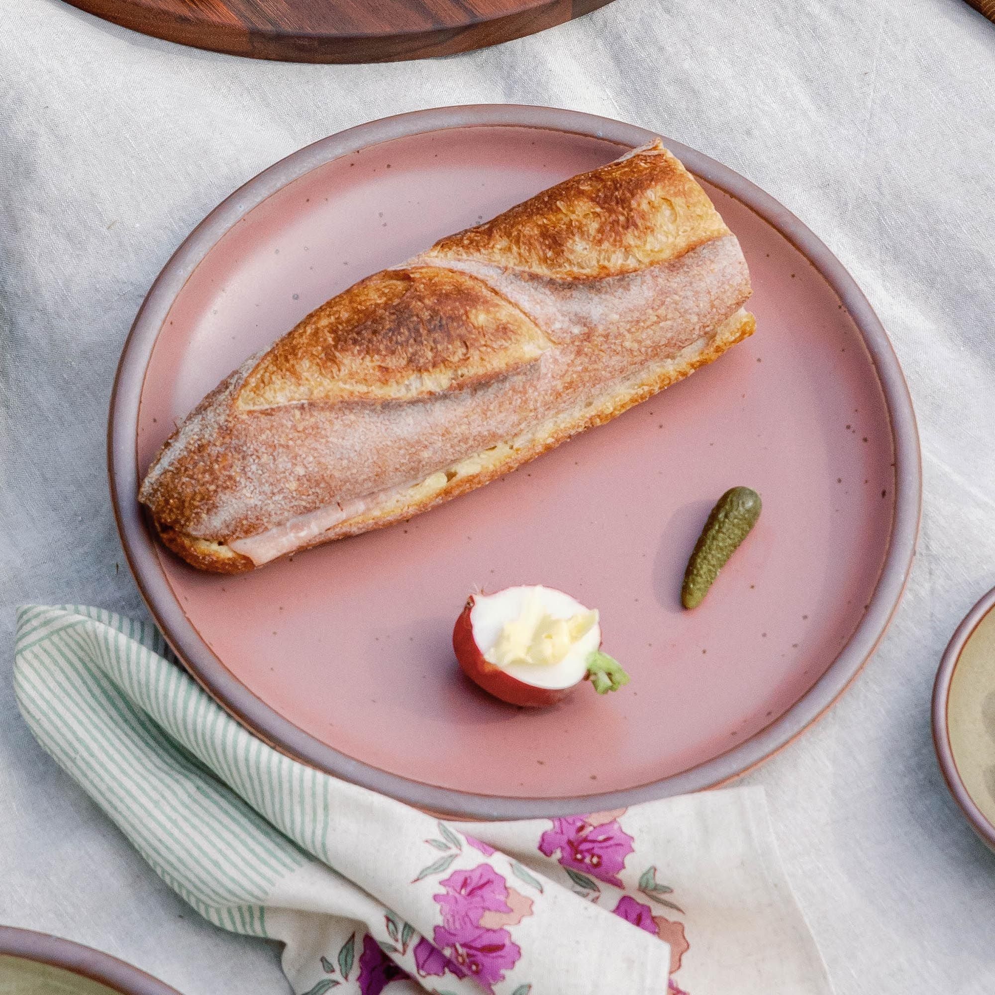 Dusty mauve pink ceramic plate with a baguette sandwich, a radish with butter, and a small pickle on a linen tablecloth.