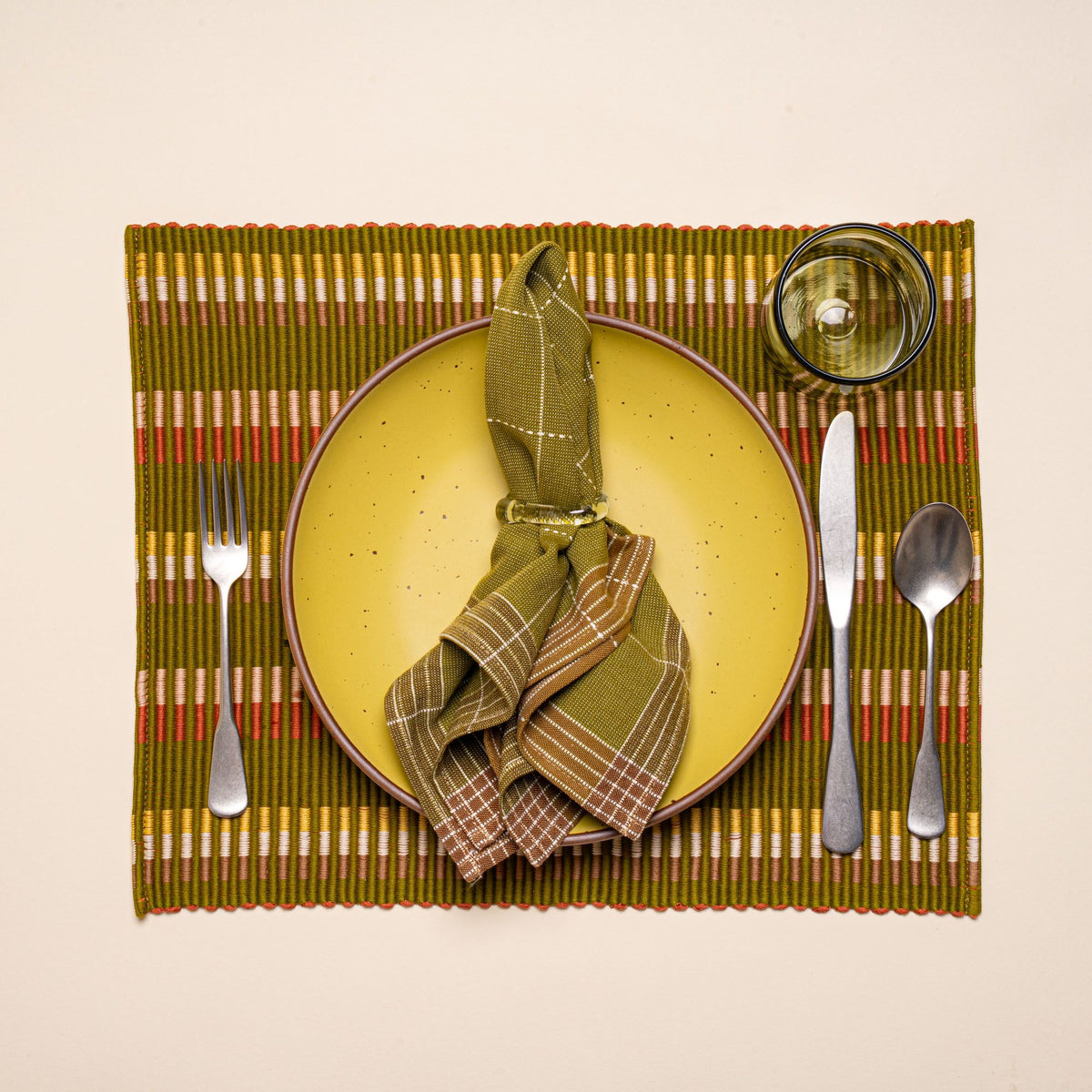 A yellow ceramic plate with silverware, glass, a green grid napkin on a rectangle placemat with offset stripe design that is hand woven in olive, tan, orange, and yellow colors