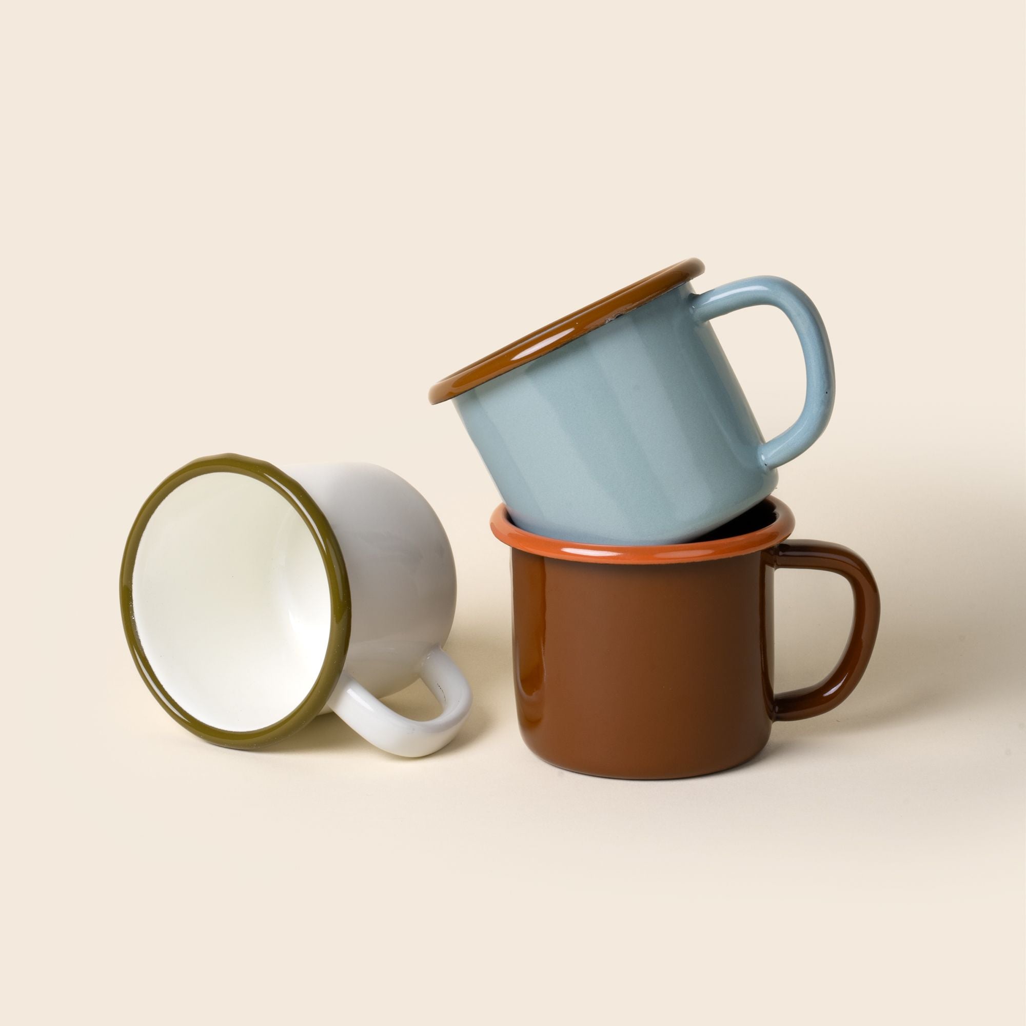Three enamel mugs - white with olive rim, blue with brown rim, and red-brown with orange rim - stacked on a beige background.