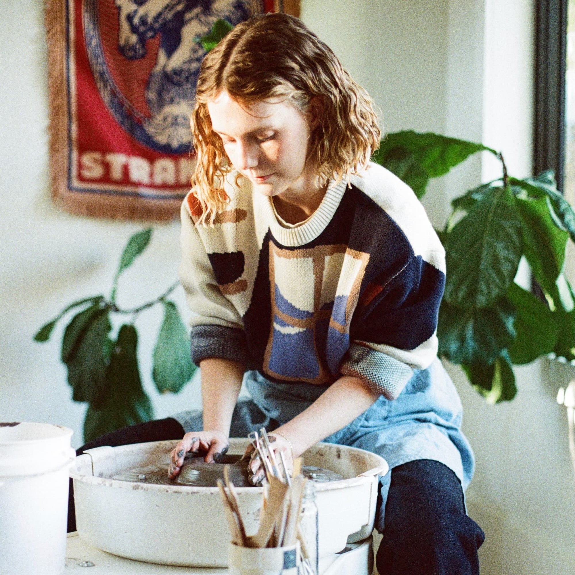 Erica is working at the potter's wheel and wears a sweater with an illustration of a pottery workshop with a large window, shelves, and table with mugs and bowls.