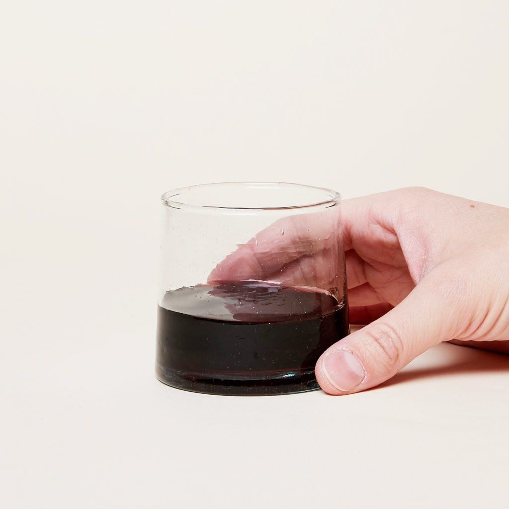 A hand holds a short clear drinking glass that contains some red wine