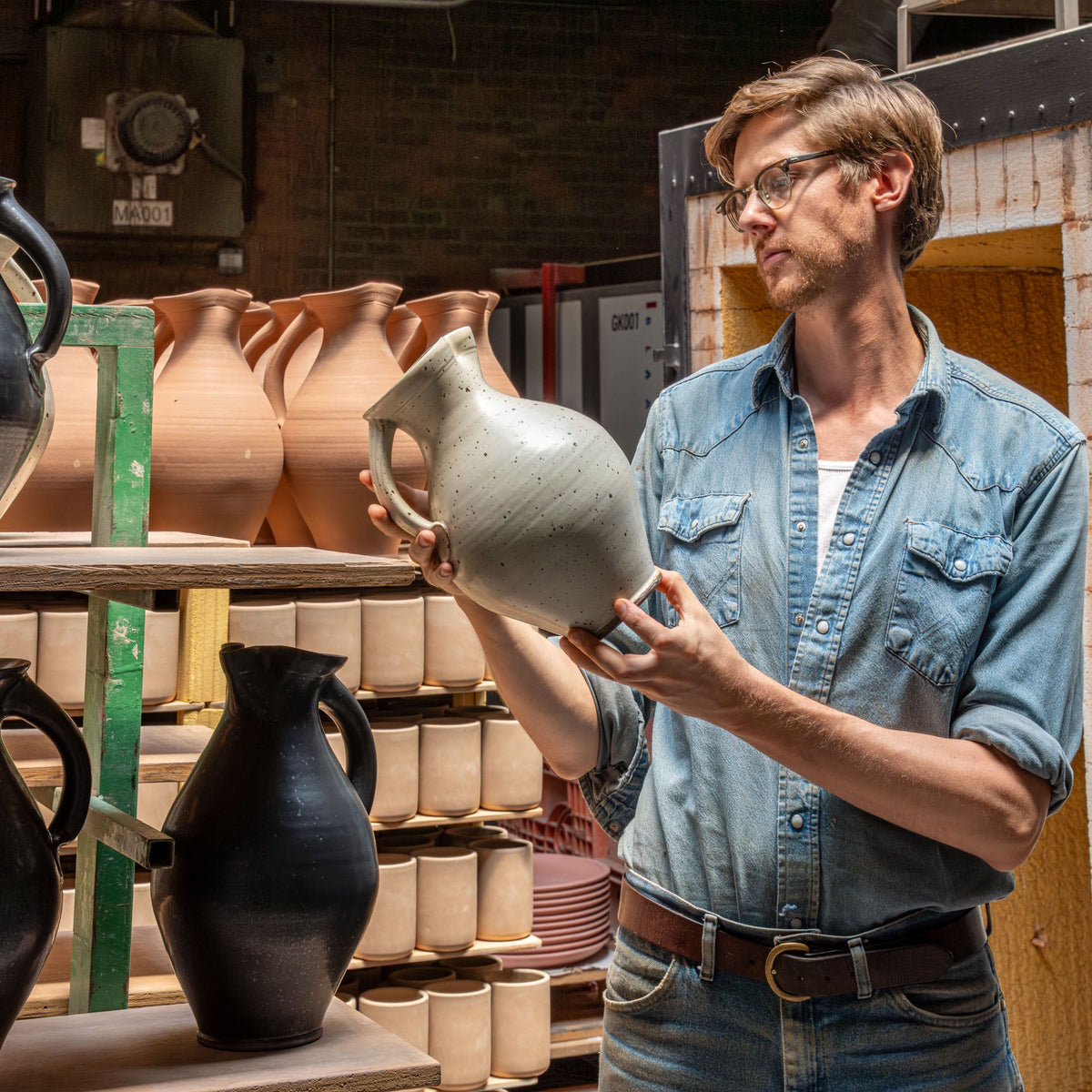 John holding a finished pitcher in the factory in front of pitchers ready to be fired in the kiln.