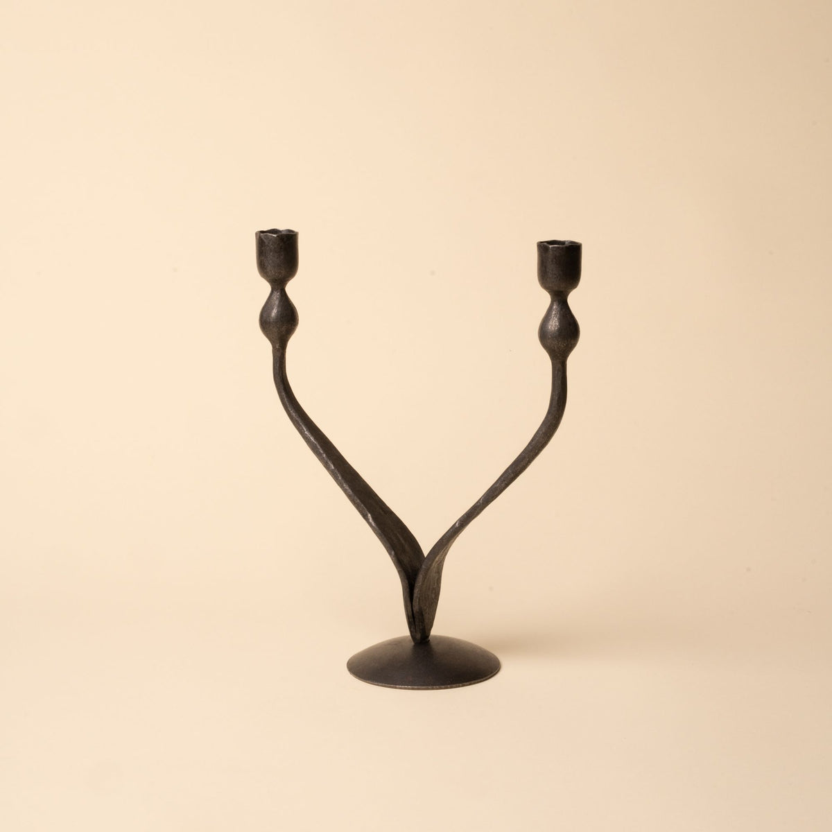 Forged iron double-arm candleholder with curved branches and round base, photographed on a beige background.