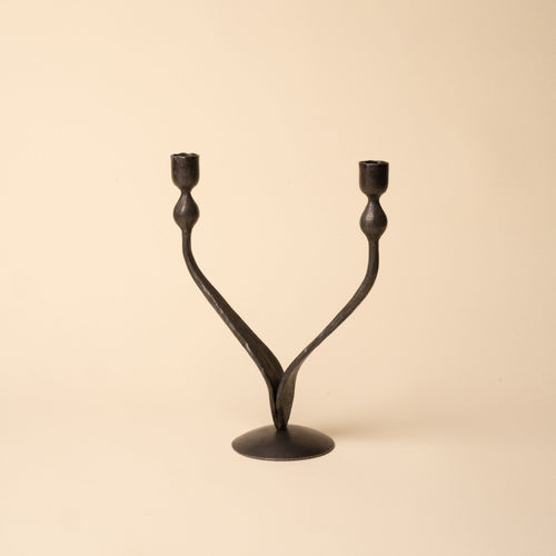 Forged iron double-arm candleholder with curved branches and round base, photographed on a beige background.