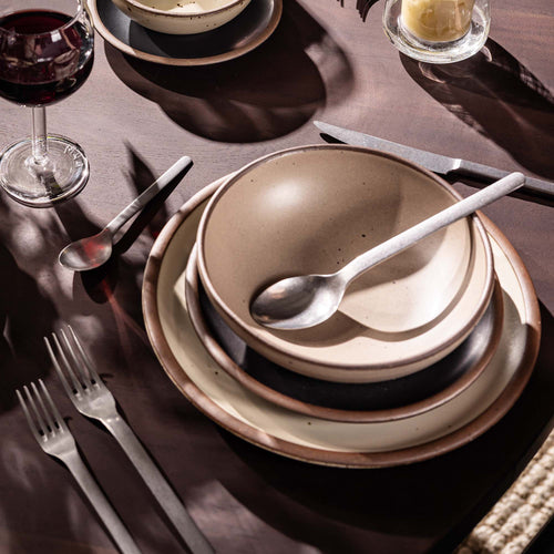 A place setting with a dinner plate, side plate, and shallow dinner bowl in natural, cream, and graphite colors, with a glass of wine and steel flatware.