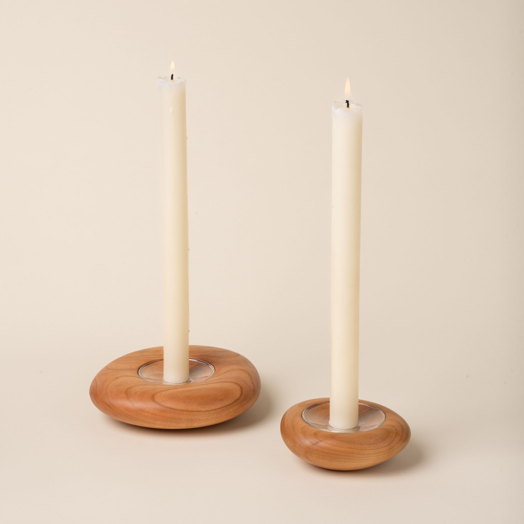 Two lit white taper candles in round, cherrt wood candle holders with visible grain, displayed against a neutral background.