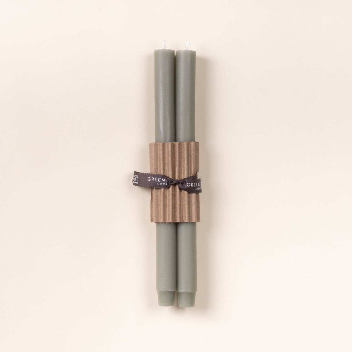 Two pale sage green taper candles bundled with corrugated cardboard and tied with a black ribbon on a neutral background.