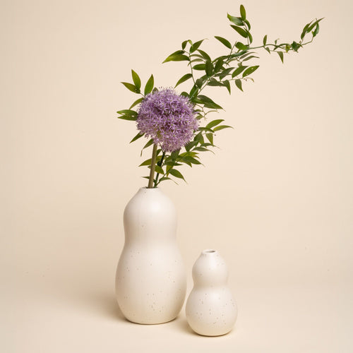 A large and small off-white speckled curvy vases, one holding a purple flower with leafy green stems, set against a neutral background.