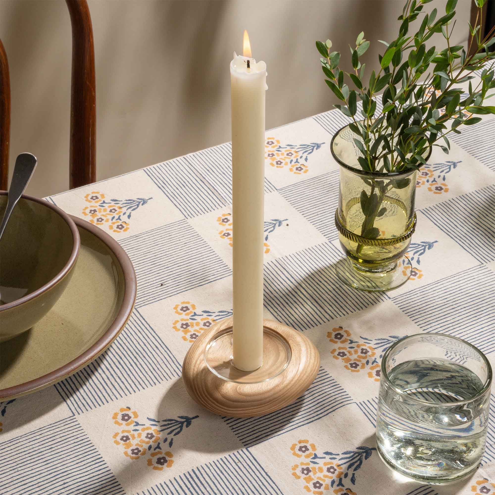 Lit white taper candle in a light wooden holder on a patterned table setting with glassware and olives.