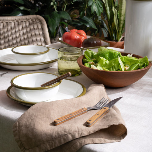 An outdoor table set with a light warm brown napkin, enamelware bowls, wood-handled cutlery, green salad, and glass of iced water, surrounded by lush plants.