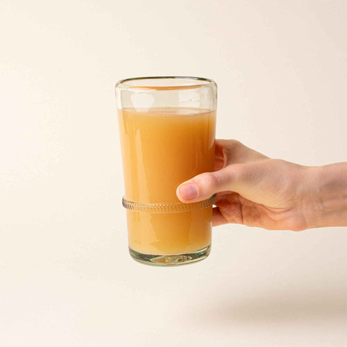 A hand holds a clear tall recycled glass tumbler with a ridged band detail, filled with iced coffee.