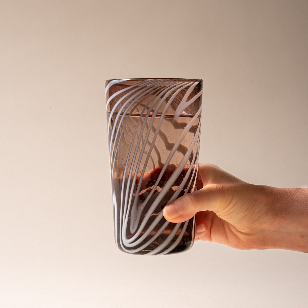 A hand holds out a smoky brown pint glass with white diagonal swirl pattern on a neutral background.