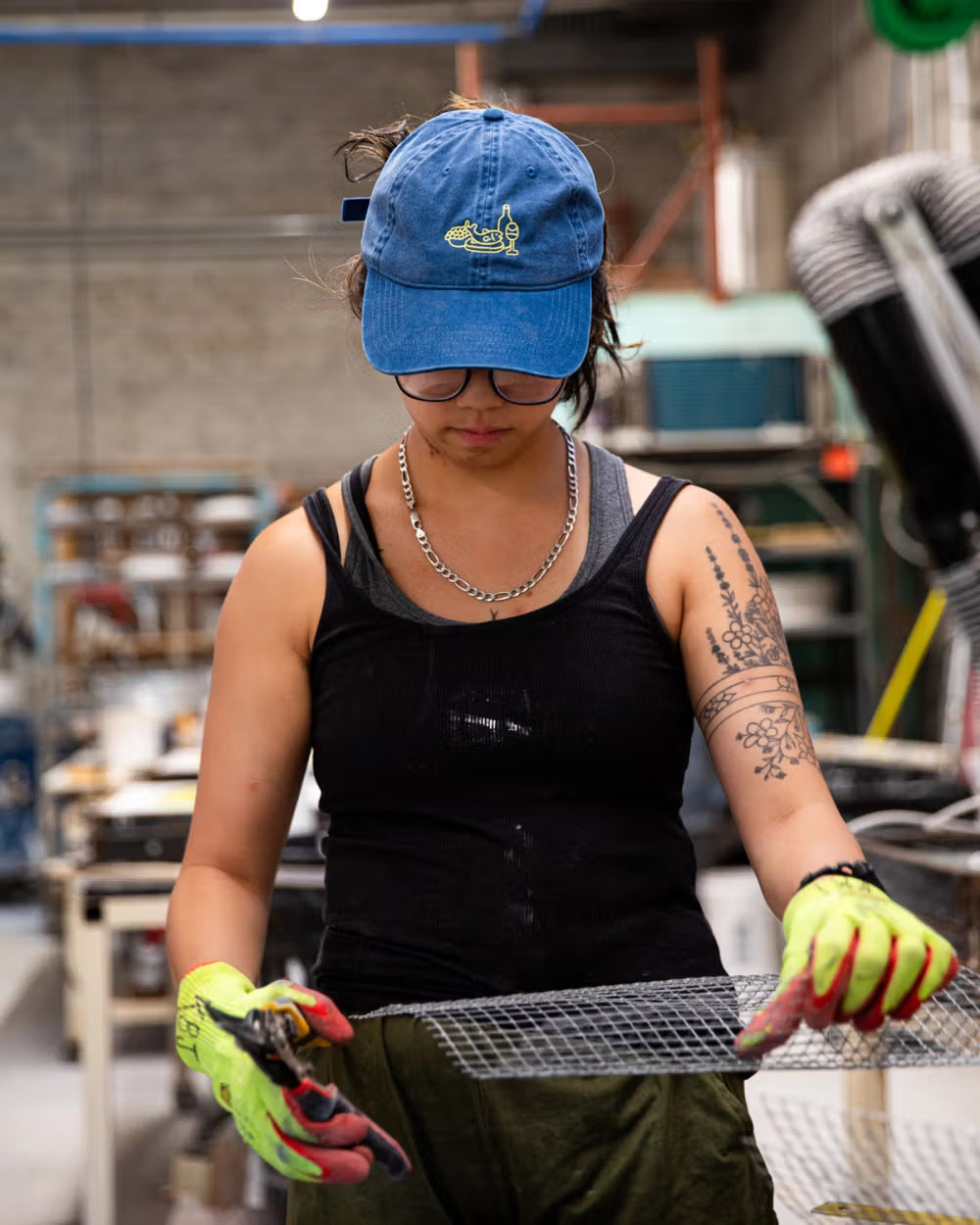 A person wearing a blue cap, glasses, and a black tank top is working with a piece of wire mesh in an industrial setting. They are holding tools and wearing bright yellow safety gloves.