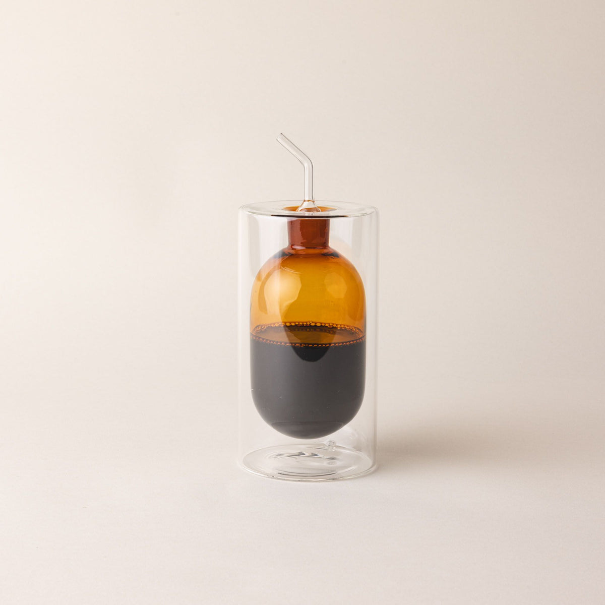 A hand holds an artful glass cruet in an orange color. There is a rounded bulb that acts as a container for the cruet, surrounded by a doubled glass wall. The cruet is filled with oil.