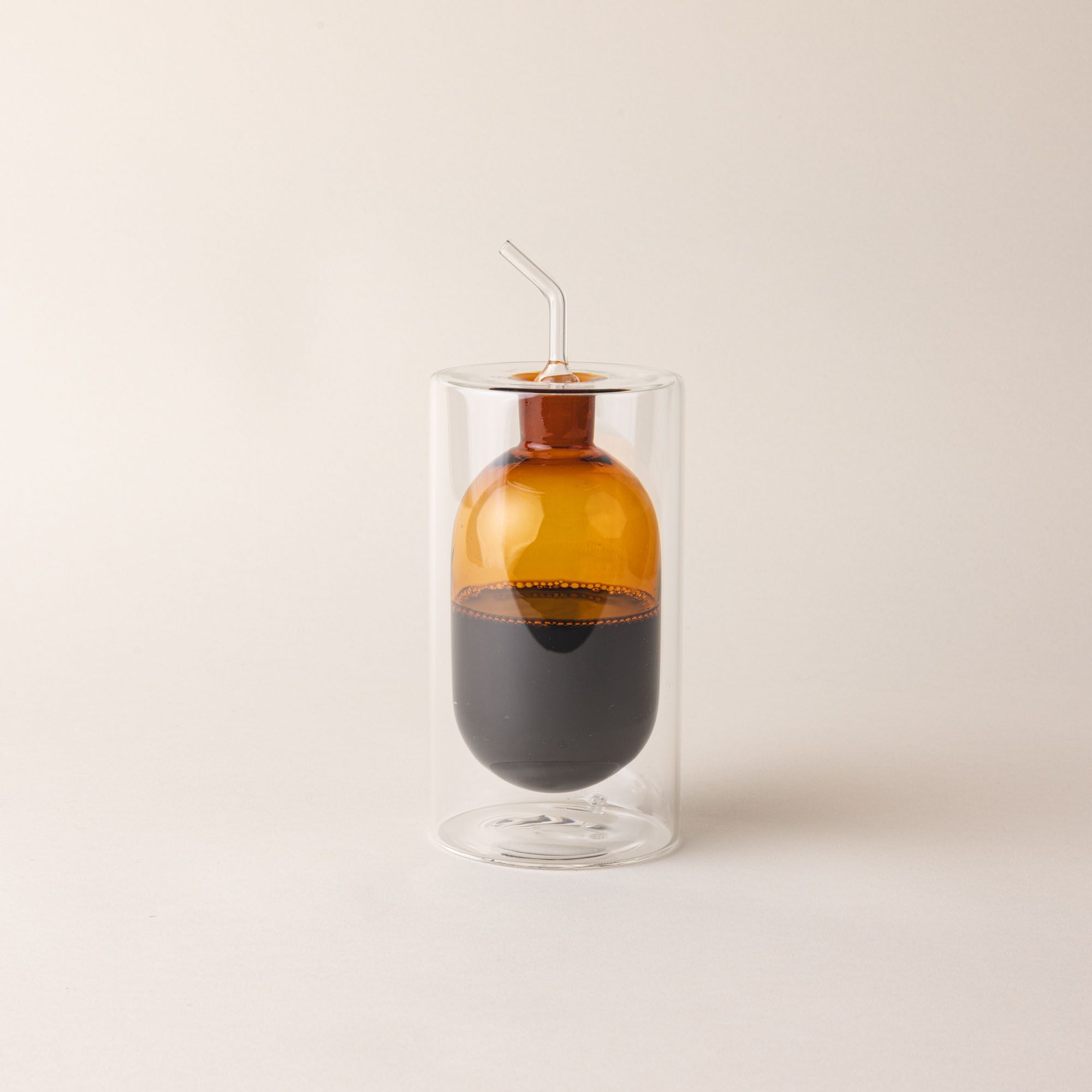 A hand holds an artful glass cruet in an orange color. There is a rounded bulb that acts as a container for the cruet, surrounded by a doubled glass wall. The cruet is filled with oil.