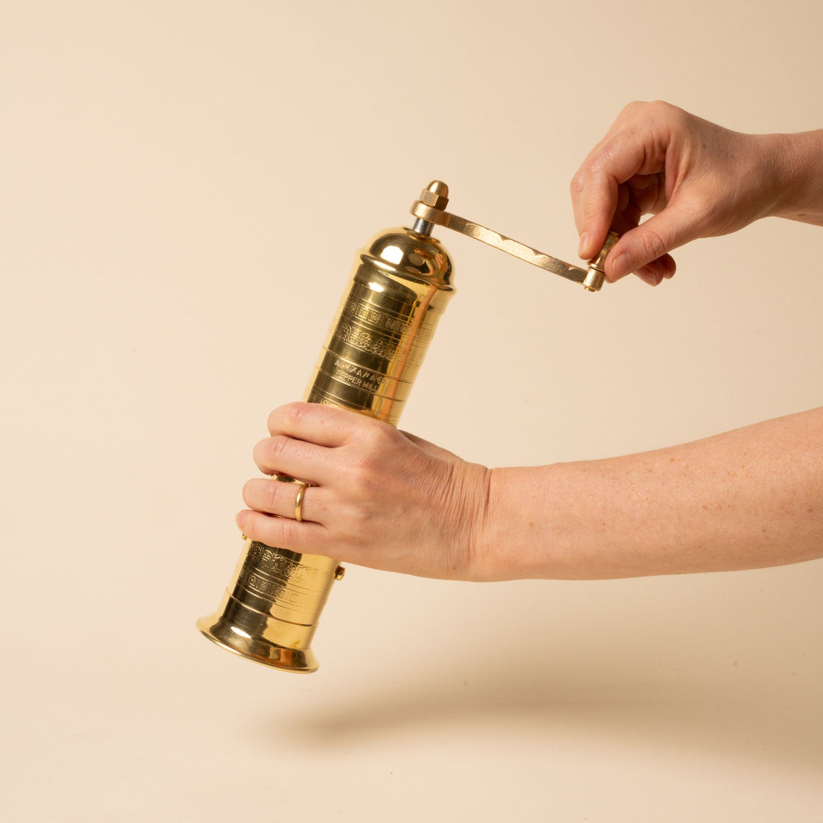 Hand holds a brass hand-crank pepper grinder with engraved details, standing upright on a neutral background.