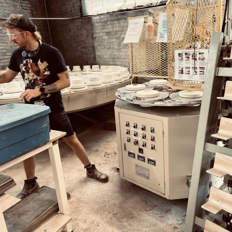 A person in a workshop stands near a pottery machine with control switches and plates in progress. Shelves with ceramic items are visible in the background, and the setting has a rustic, industrial feel.