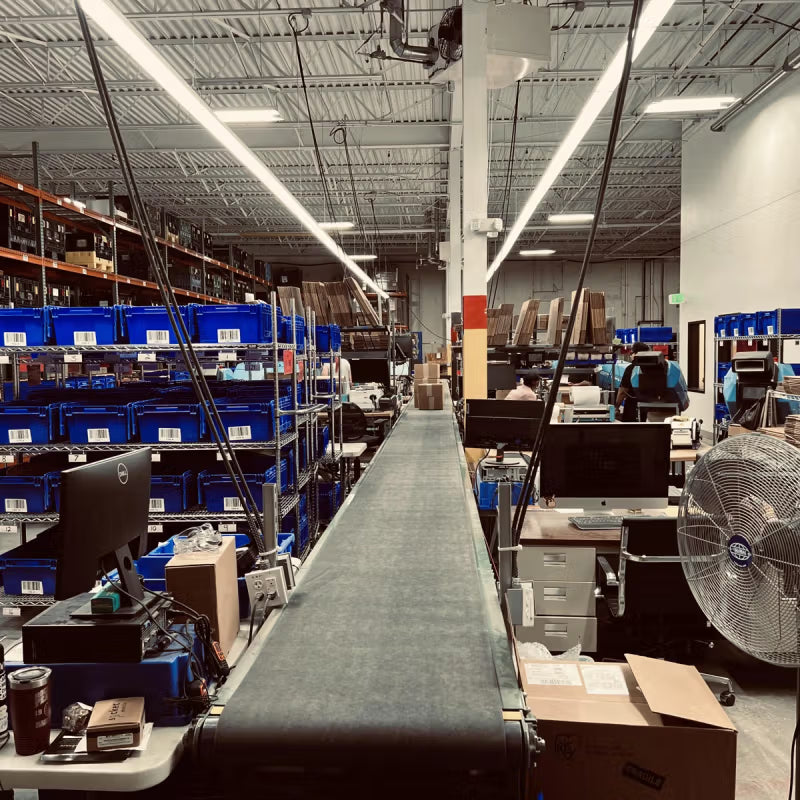 Interior of a fulfillment center with a conveyor belt running through the middle. Blue bins are stacked on shelves alongside the belt, and workstations with computers and packaging materials are set up nearby.