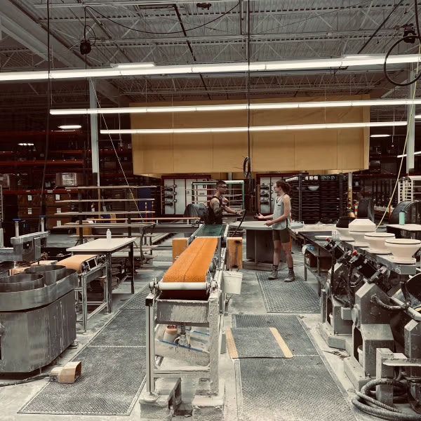 Two people in a ceramics workshop are engaged in the glazing and firing process, surrounded by pottery equipment, tables, and shelves. The setting is industrial with visible overhead lighting and unfinished pottery pieces.