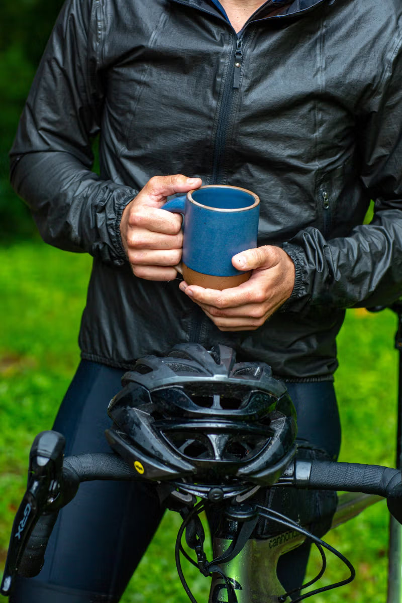 A person in a black jacket stands outdoors, holding a blue mug, with a bicycle helmet resting on the handlebars of a bike.
