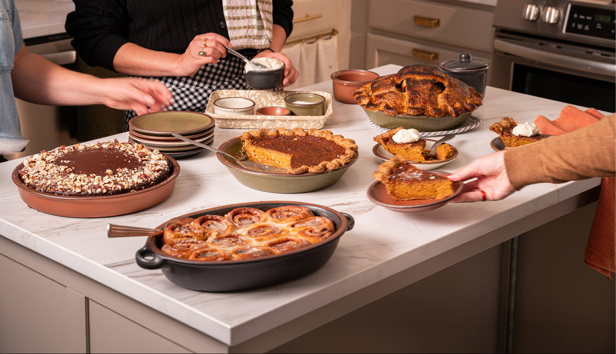 A kitchen scene with multiple baked goods on a countertop, including pies, cakes, and cinnamon rolls. Several people are visible from the shoulders down, serving and holding food items.