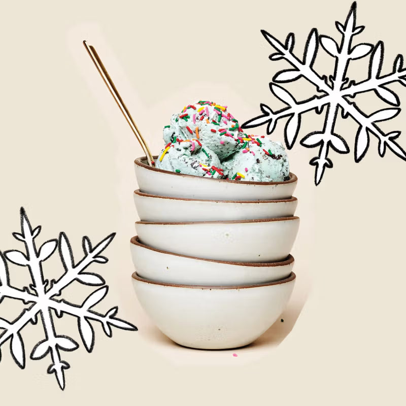 A stack of five cream-colored ceramic bowls, with the top bowl filled with mint chocolate chip ice cream and sprinkles, and a brass spoon beside it. Two large snowflake illustrations decorate the background.