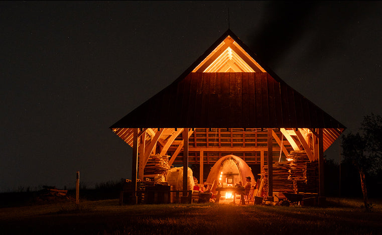 A cabin at night with a wood fired kiln firing with the whole cabin aglow and smoke coming out of the top.