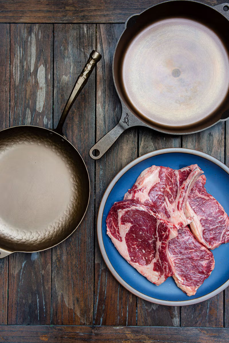 Two metal pans and a plate of raw steaks on a rustic wooden surface.