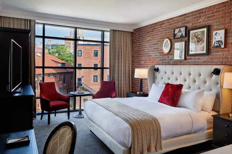 Hotel room with a brick accent wall, featuring a large bed with a tufted headboard, white bedding, and a red throw pillow. Two red chairs and a small round table are positioned by the window, with framed artwork on the wall and a view of a building outside.