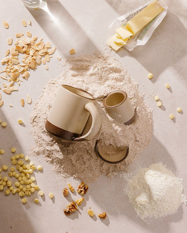 Two ceramic mugs, one larger cream mug with an unglazed base and one smaller matching beige mug, arranged on a mound of flour with scattered oats, walnuts, and a small bowl of flour on a warm beige surface.