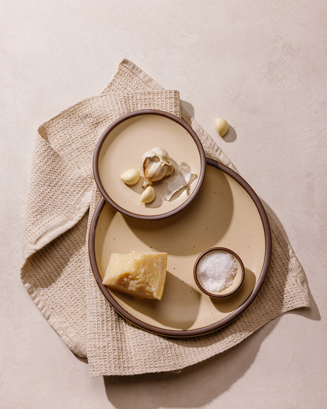Two round ceramic plates in a cream color with unglazed rims, styled with food including garlic, parmesan cheese, and salt on a beige textured cloth over a neutral background.