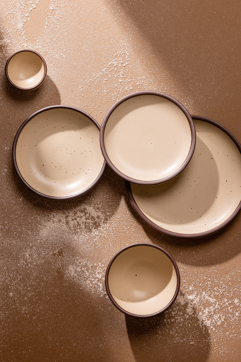 A set of ceramic tableware including plates and bowls in a cream color with unglazed rims, arranged on a textured brown surface with a light dusting of flour.