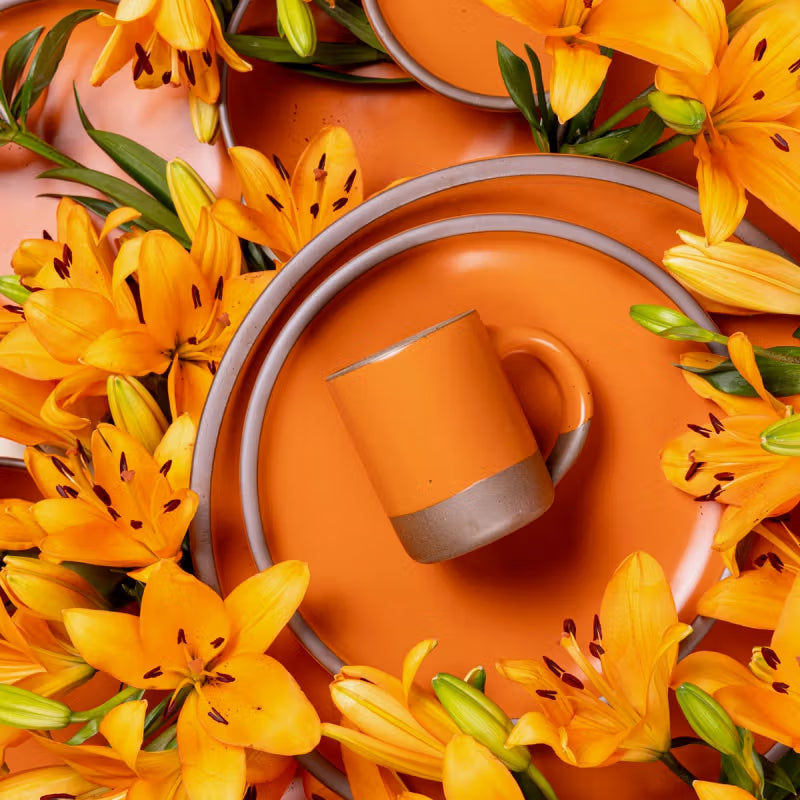 Orange ceramic plates and a mug in the "Daylily" glaze are surrounded by vibrant orange lilies with green leaves.