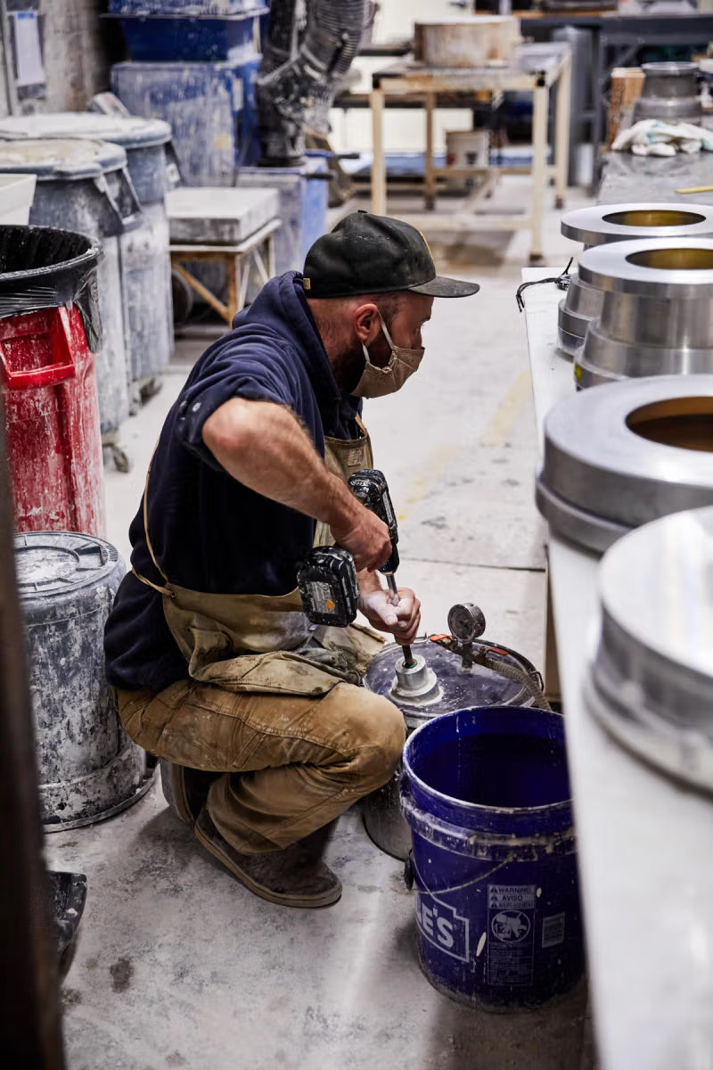 A person wearing a cap and mask is kneeling in a pottery mold shop, operating machinery. The workshop is filled with various containers and equipment, creating a busy industrial setting.